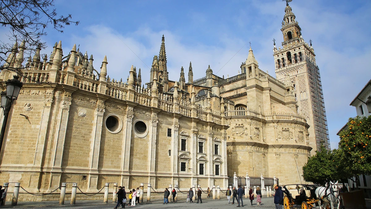 Gothic cathedral with a tall bell tower, people and a horse carriage under a clear sky.
